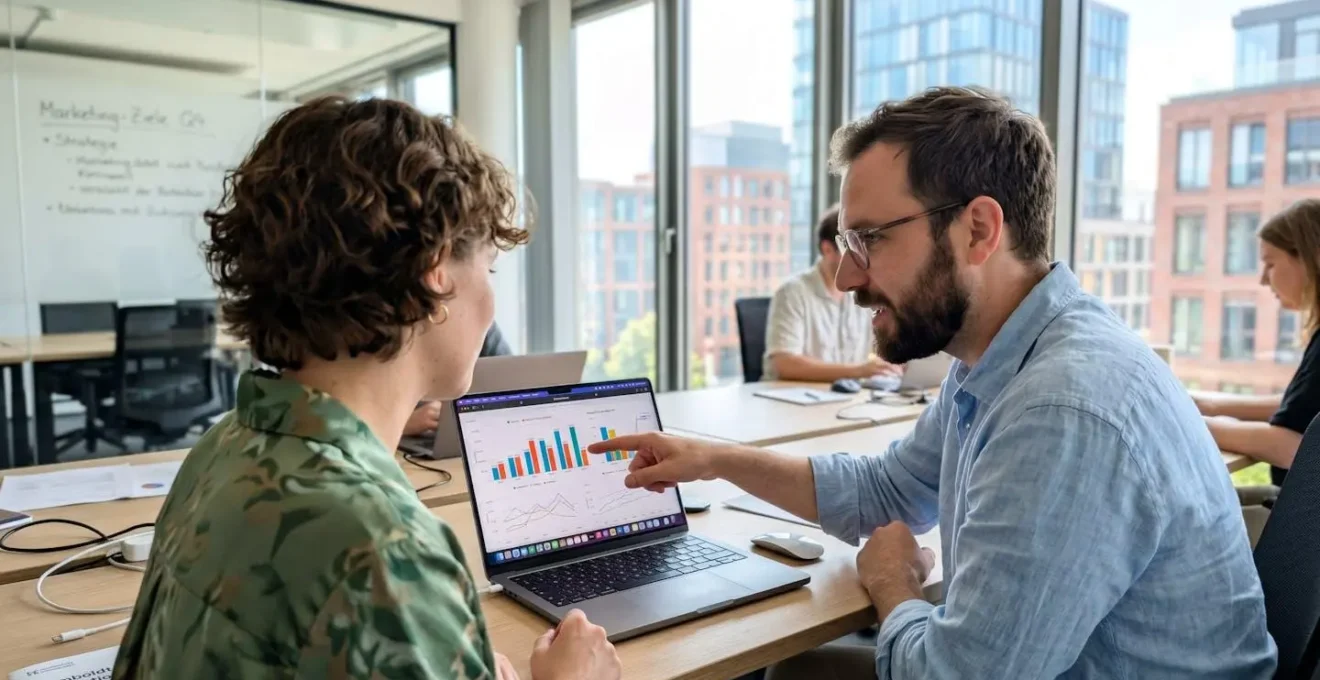 Zwei Marketing-Fachleute betrachten gemeinsam farbige Diagramme und Grafiken auf einem Laptop-Bildschirm in einem hellen modernen Büro